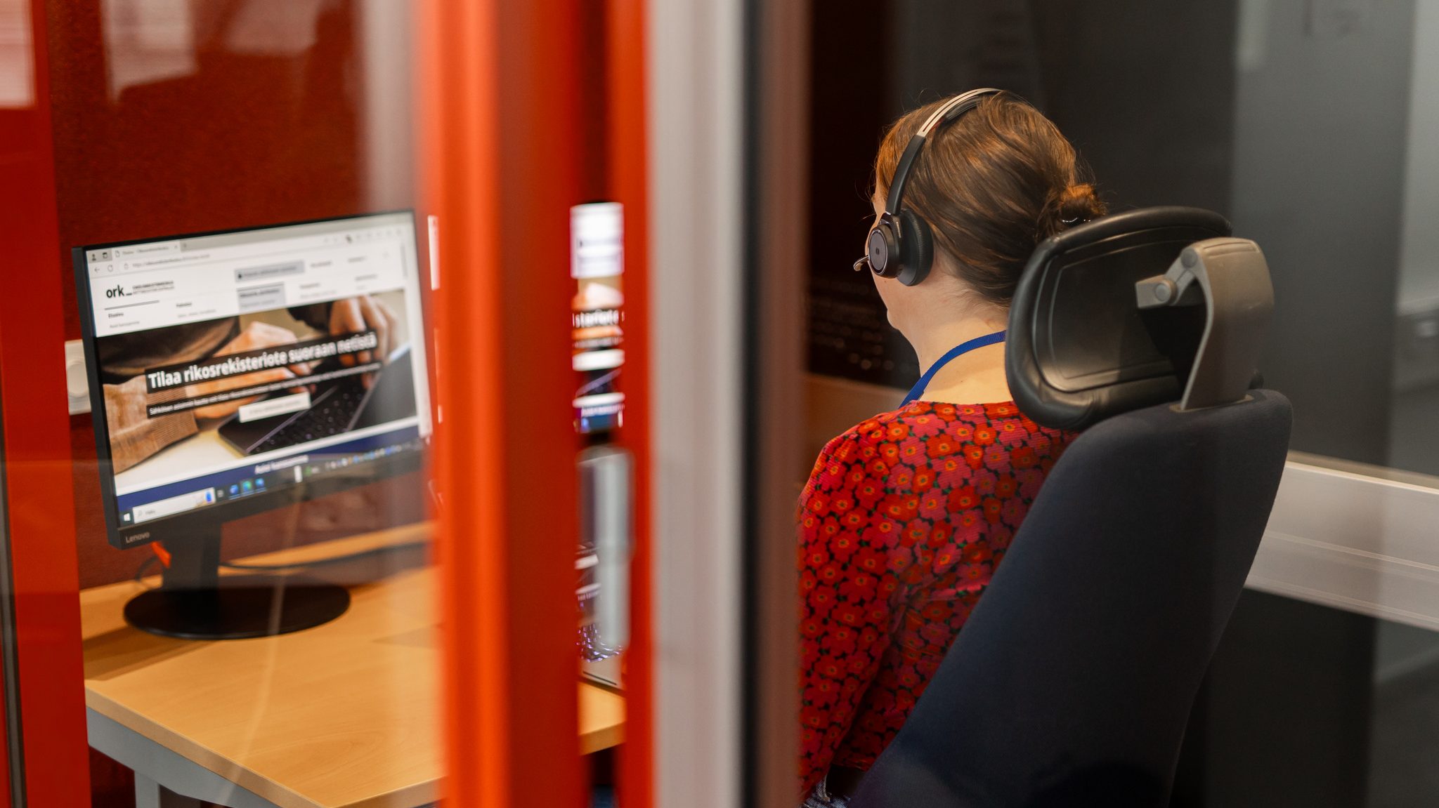 En person sitter på en kontorsstol och använder en dator med hörlurar på huvudet. På skärmen står det "Beställ ett utdrag ur straffregistret direkt från nätet".