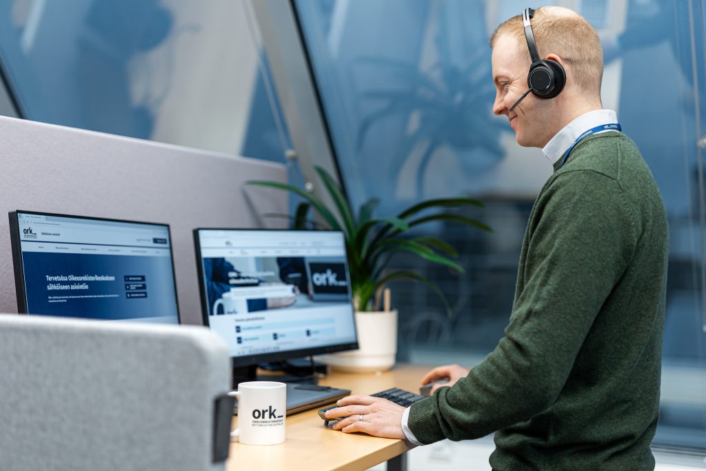 Mieshenkilö tekee töitä työpöydän ääressä kuulokkeet päässä. Työpöydällä on kaksi tietokoneen näyttöä, joissa toisessa näkyy ORK:n verkkosivut ja toisessa ORK:n sähköinen asiointi.
