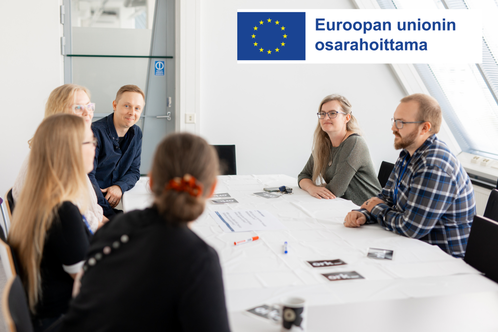 Pöydän ääressä istuu kuusi ihmistä. Pöydällä näkyy erilaisia papereita. Kuvan oikeassa yläkulmassa on EU:n logo ja teksti "Euroopan unionin osarahoittama".
