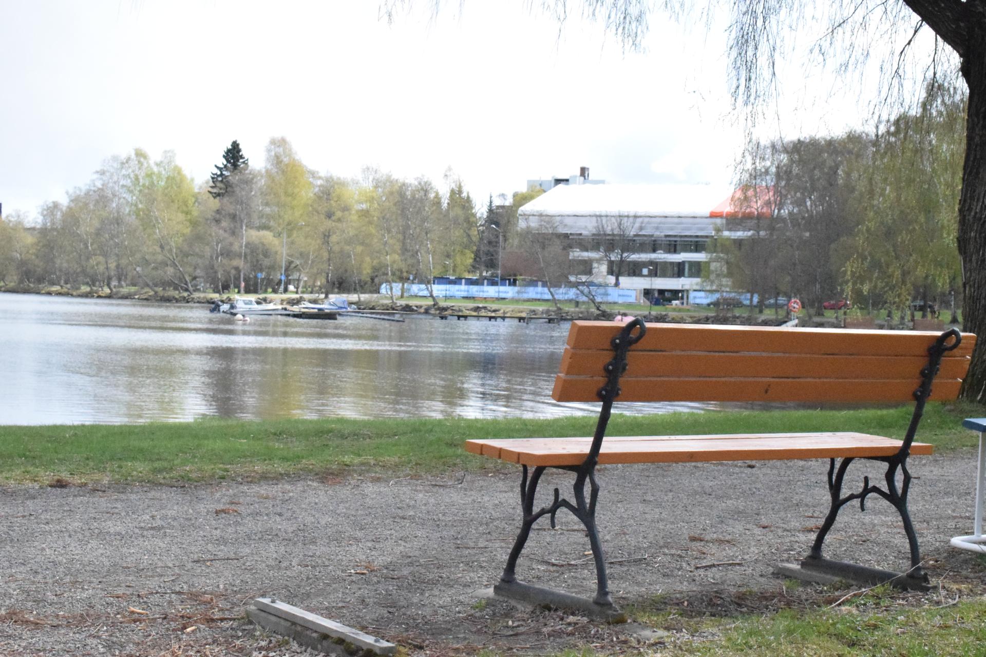 Taustalla virtaa joki ja etualla on kävelytie ja puiston penkki Taustalla virtaa joki ja etualla on kävelytie ja puiston penkki